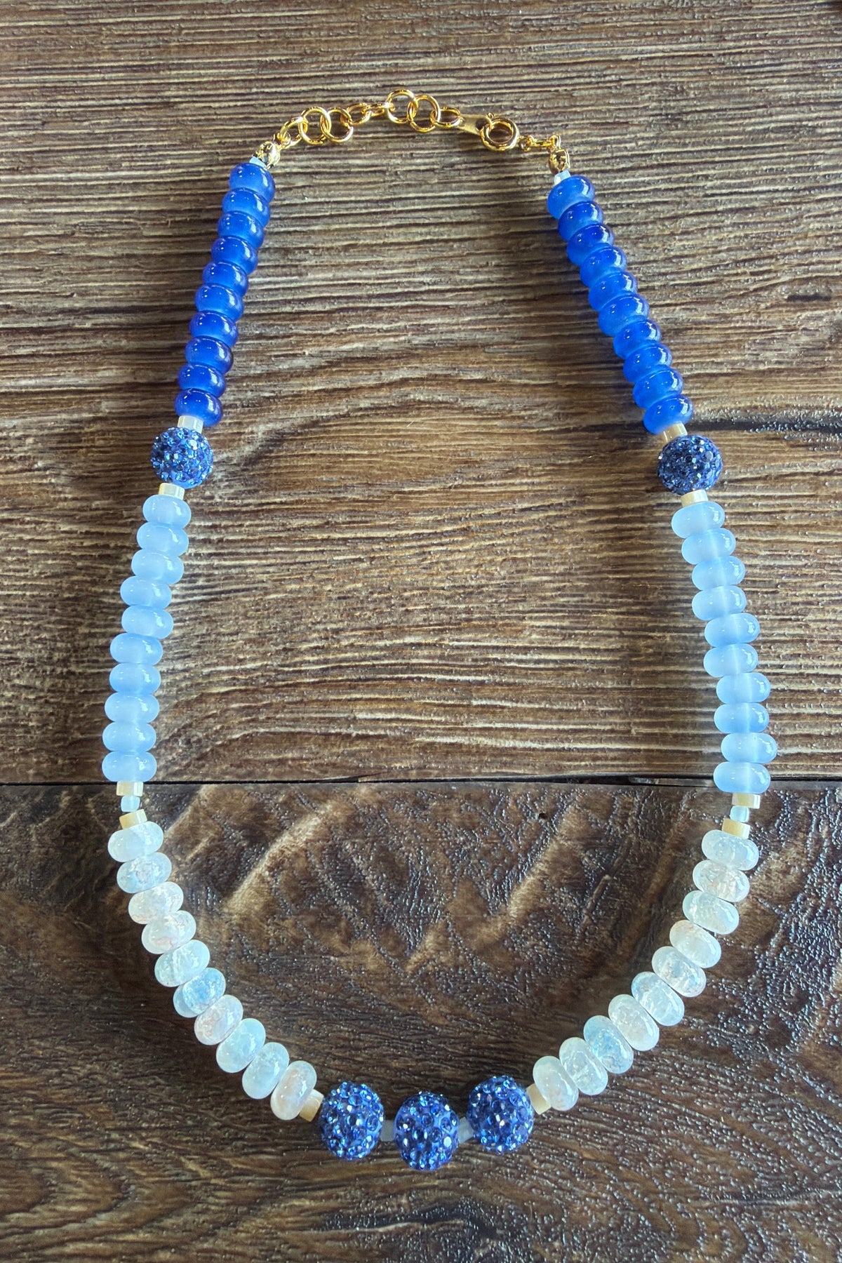 A beaded necklace featuring light blue and white faceted beads, blue textured round beads, and a gold chain clasp, displayed on a dark wood background.