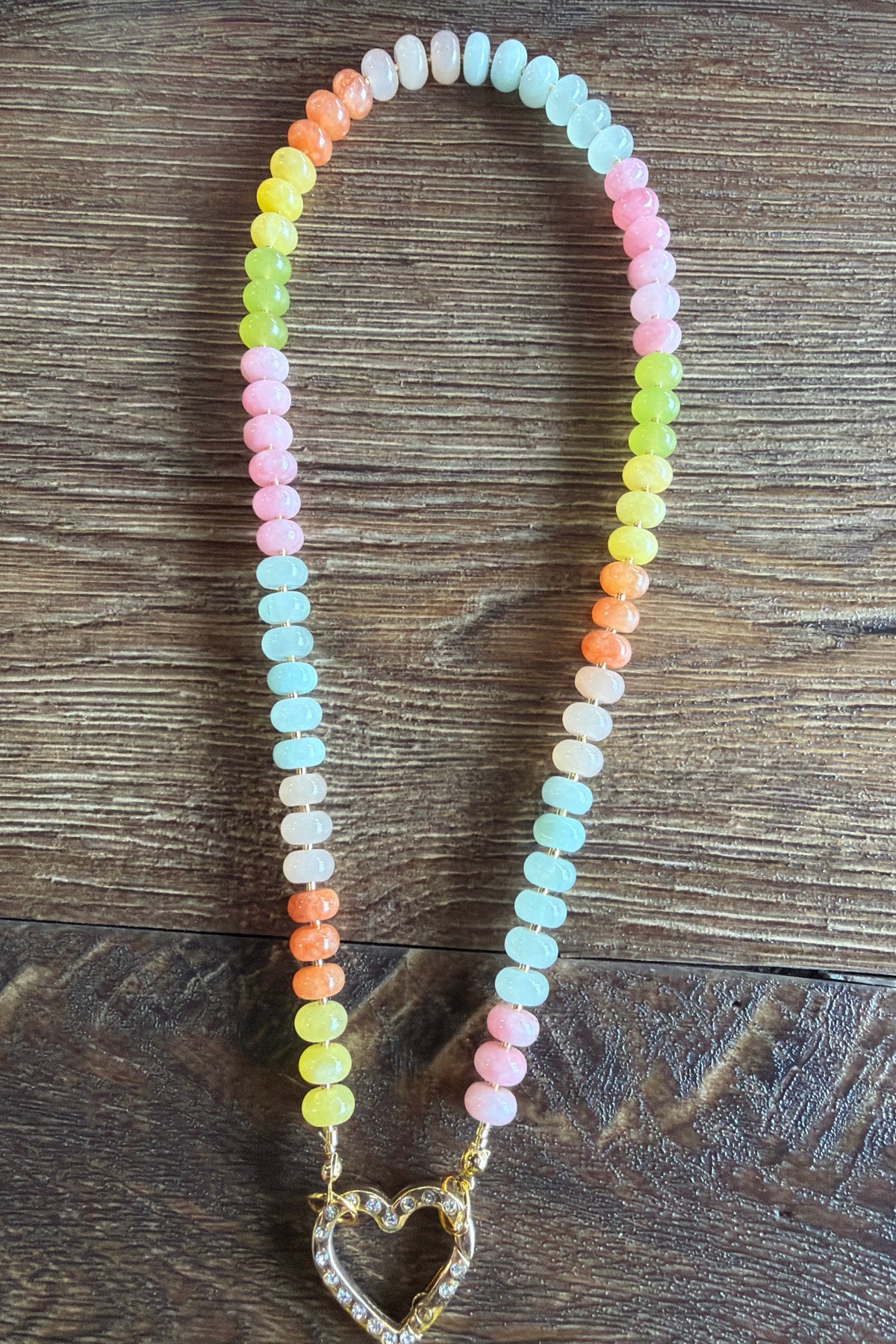 A pastel beaded necklace with a gold heart-shaped pendant, laid flat on a dark wood background. The necklace features multiple strands of round beads in pink, light blue, orange, yellow, and white, with a gold heart pendant encrusted with clear stones.