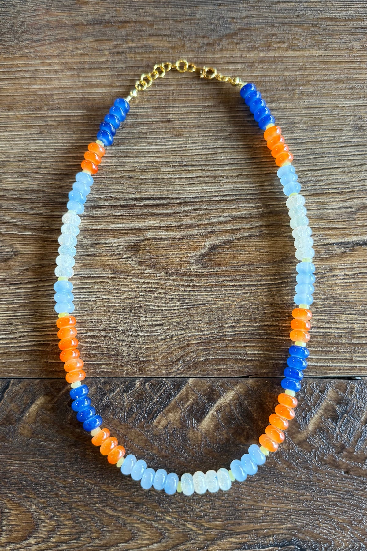 A beaded necklace with alternating rows of translucent light blue, bright blue, orange, and white beads, finished with a gold clasp, displayed on a dark wood background.