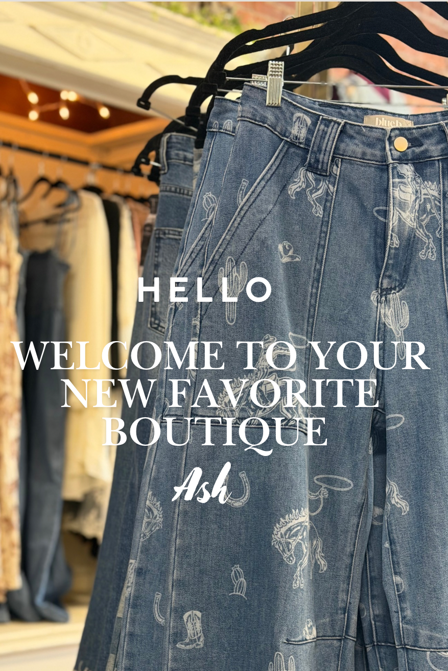 A pair of blue denim jeans with white distressed graphics hanging on black hangers in a boutique setting. White text overlay reads 'HELLO WELCOME TO YOUR NEW FAVORITE BOUTIQUE' with a script logo 'Ash'.