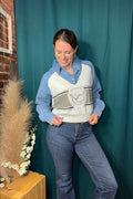 A woman modeling a light blue and white raglan sweater vest with a gray graphic print, worn over a light blue button-down shirt and paired with blue jeans, standing in front of a teal fabric backdrop.