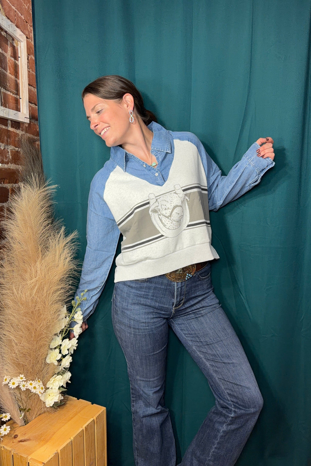 A woman posing in a light blue and white long-sleeve knit sweater vest with a gray graphic print, worn over a light blue button-down shirt and blue jeans, set against a dark teal fabric backdrop.