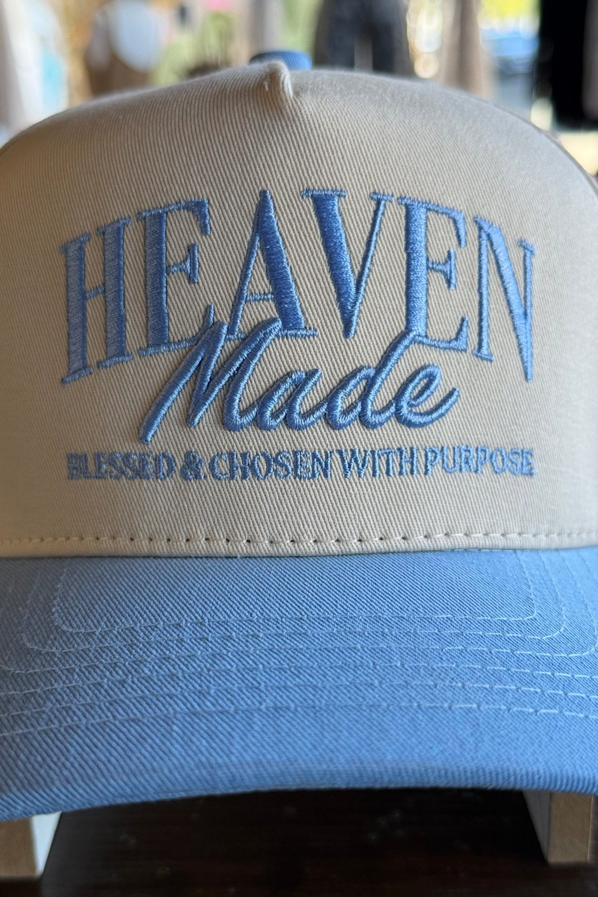 A beige and light blue trucker-style cap with 'HEAVEN Made' embroidered in blue on the front, resting on a dark wooden surface with a blurred outdoor background.