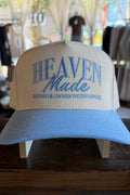 A light blue and cream snapback cap with 'HEAVEN Made' embroidered on the front, displayed on a wooden stand inside a store.