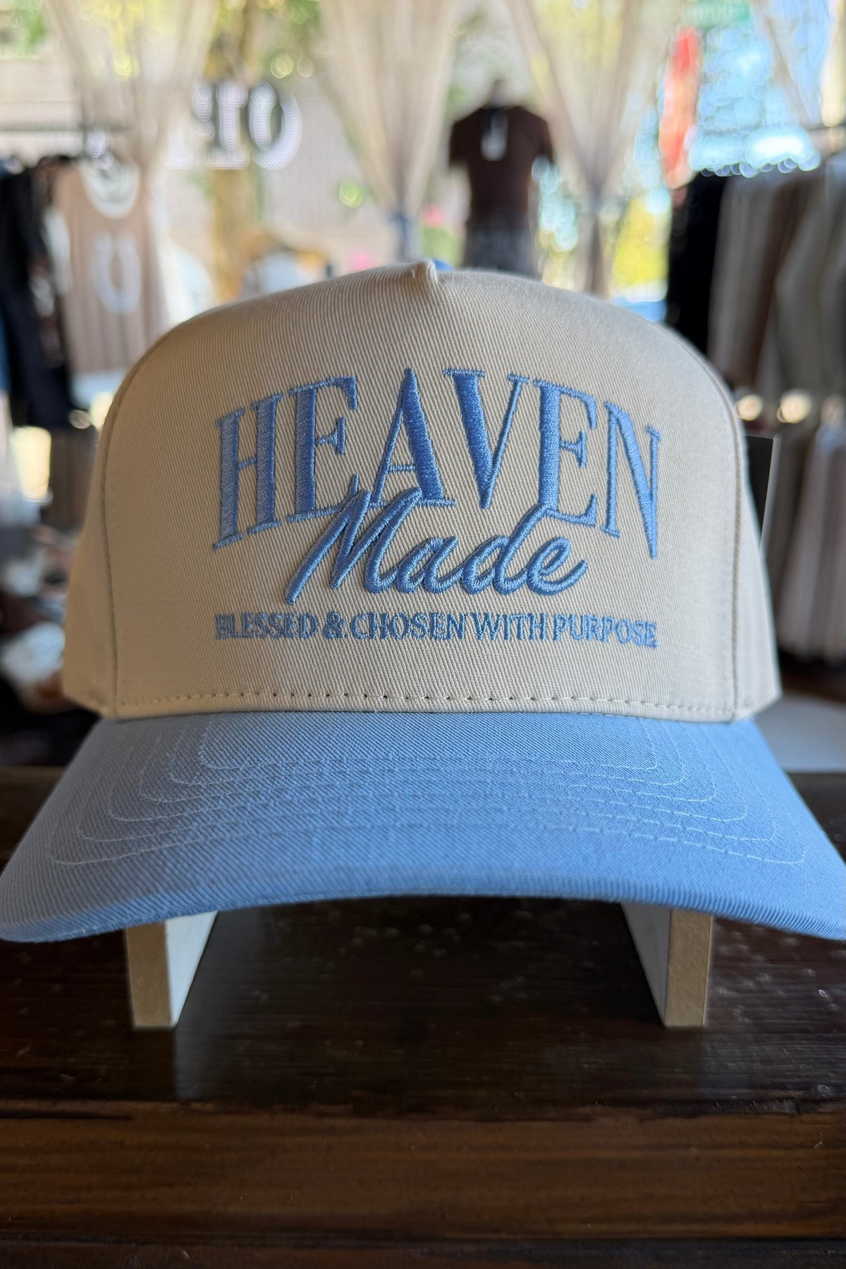 A light blue and cream snapback cap with 'HEAVEN Made' embroidered on the front, displayed on a wooden stand inside a store.