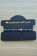 A handmade beaded bracelet with a small silver cross charm, displayed on a black card that reads 'Handmade with Love' on a light wood background.