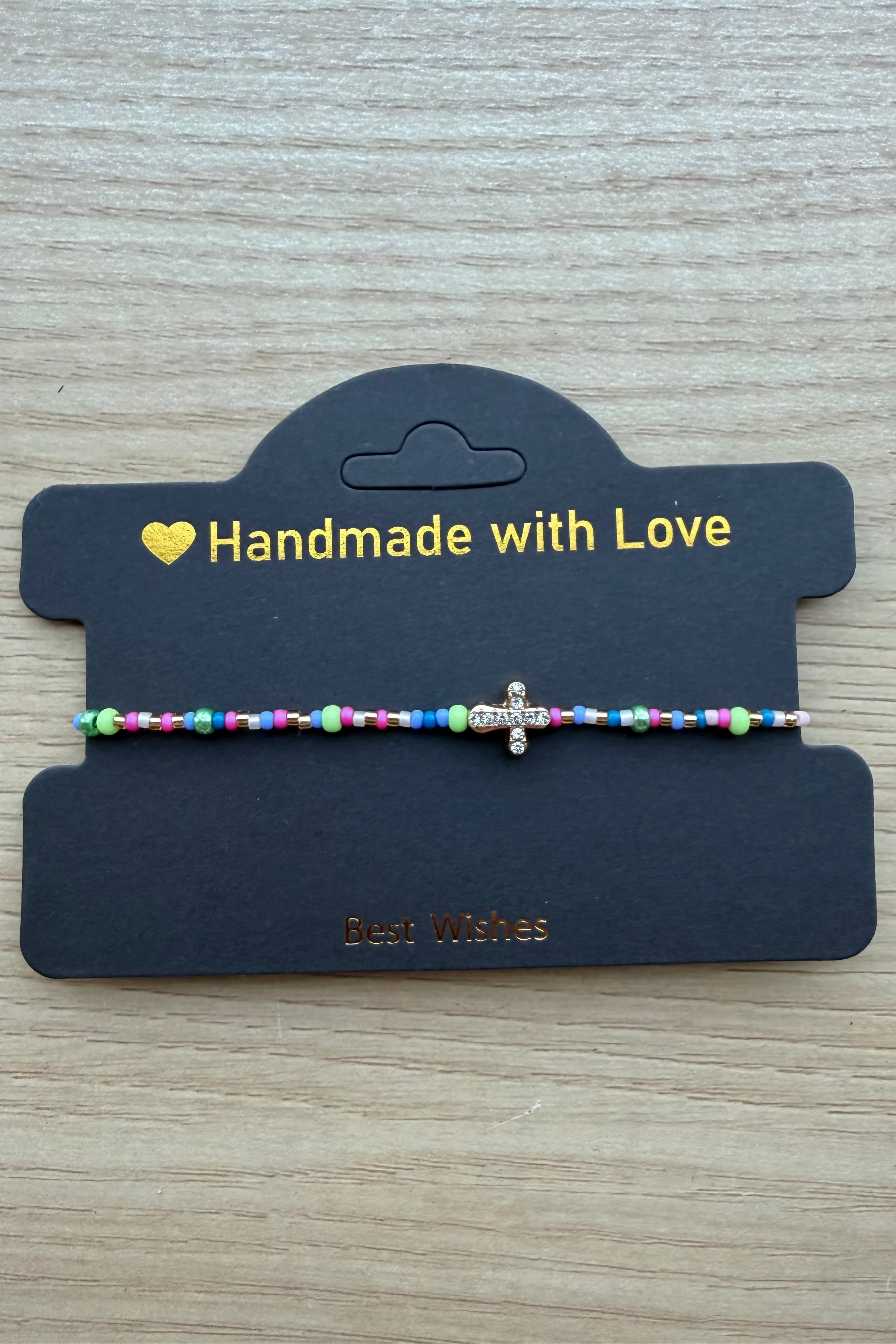 A handmade beaded bracelet with a small silver cross charm, displayed on a black card that reads 'Handmade with Love' on a light wood background.