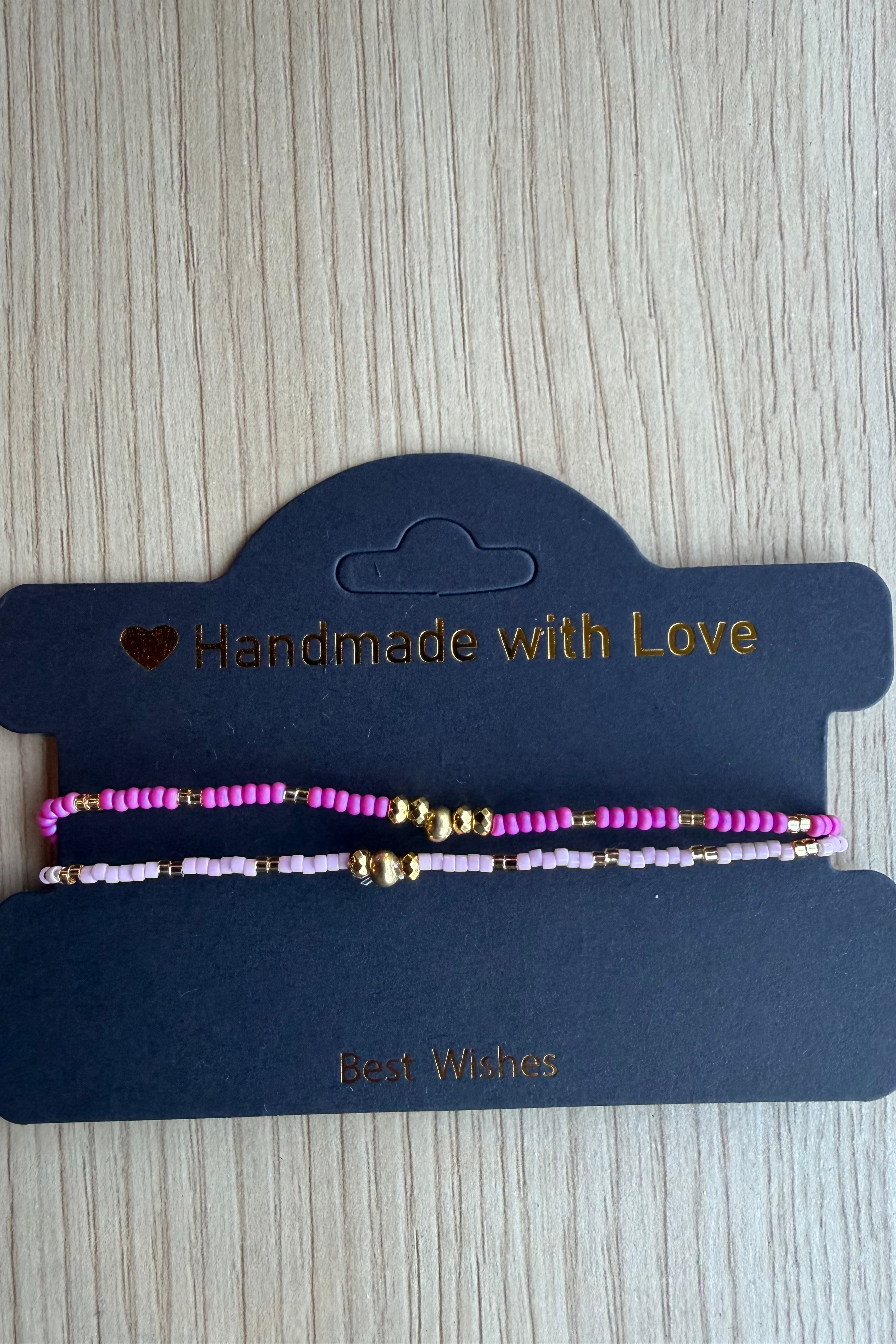 Two beaded friendship bracelets in pink and lavender with gold spacer beads, displayed on a dark blue card that reads 'Handmade with Love' and 'Best Wishes' on a light wood background.